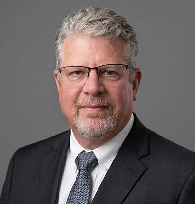 Professional corporate headshot of a middle-aged man wearing glasses, suit, and tie on a gray background – snagrp.com